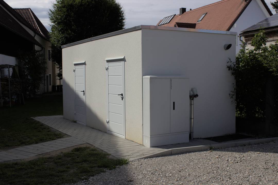 glasfaserausbau-bayern-verteilerbau-aresing-03 Glasfaserausbau Bayern: Fertig installierte Glasfaser-Verteilerstation in Aresing mit Technikgebäude und Wohngebäuden im Hintergrund